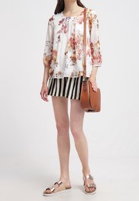 Blusa floreale leggera con maniche a tre quarti, abbinata a pantaloni corti a righe bianche e nere. Borsa a tracolla marrone e sandali metallici.