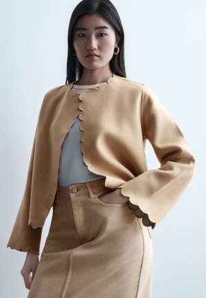 Tânără cu păr lung și întunecat, purtând o jachetă bej cu margini ondulate și o fustă asortată, pozând cu o mână în buzunar pe un fundal simplu.