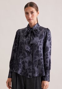 Blusa a maniche lunghe in satin grigio scuro con motivo a stampa leopardata, dotata di colletto a fiocco e maniche a palloncino arricciate.