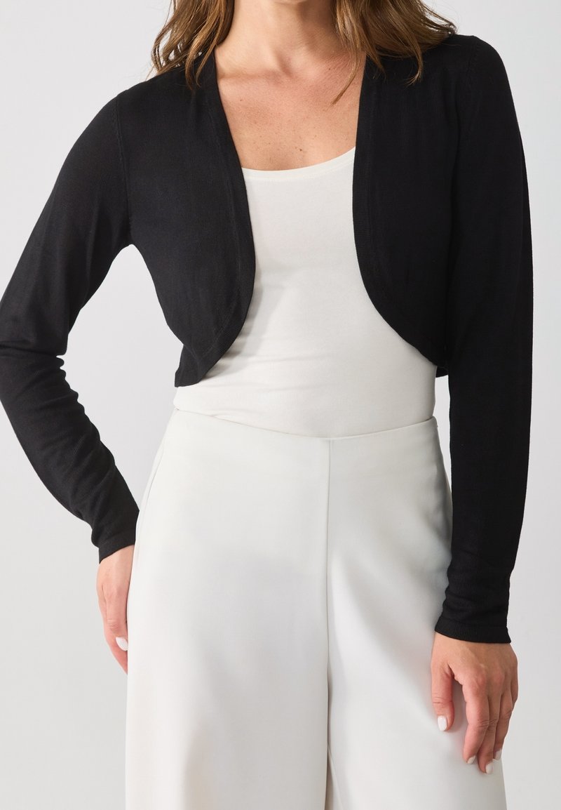 Femme portant un cardigan court noir par-dessus un haut blanc et un pantalon blanc taille haute, une main posée sur la hanche.