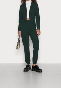 Zelený krátký blazer a odpovídající cargo kalhoty, doplněné černými šněrovacími botami. Outfit má hladkou látku a elastické manžety.