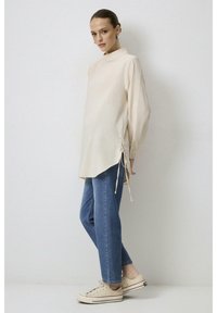 Haut beige à manches longues avec un col montant et des liens sur les côtés, associé à un jean bleu et des baskets beiges. Tissu lisse et coupe décontractée.