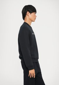 Zwarte sweatshirt met ribgebreide boorden en onderkant, met subtiele branding op de borst. Zachte textuur; ontworpen voor comfort. Zijaanzicht.