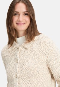 Gilet texturé de couleur crème avec un col, fermeture par boutons sur le devant et un matériau doux et duveteux.