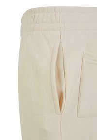 Pantalon de jogging beige en tissu texturé, avec une taille élastique, des poches latérales et une poche arrière simple avec des détails de couture visibles.