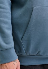 Μπλε φούτερ από απαλό ύφασμα, με μπροστινή τσέπη, ριμπ μανσέτες και τελείωμα. Η υφή είναι λεία με ελάχιστες εμφανείς ραφές.