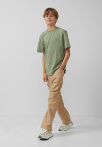 Groene katoenen T-shirt, ontspannen pasvorm, gecombineerd met beige broek. Sneakers met een textuur in grijs en crème ontwerp. Eenvoudige achtergrond.