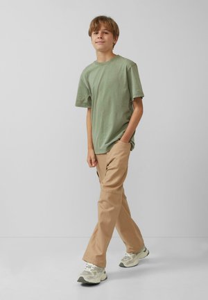 Grünes Baumwoll-T-Shirt, lässige Passform, kombiniert mit beigen Hosen. Dazu Sneaker mit einem strukturierten Design in Grau und Creme. Einfacher Hintergrund.