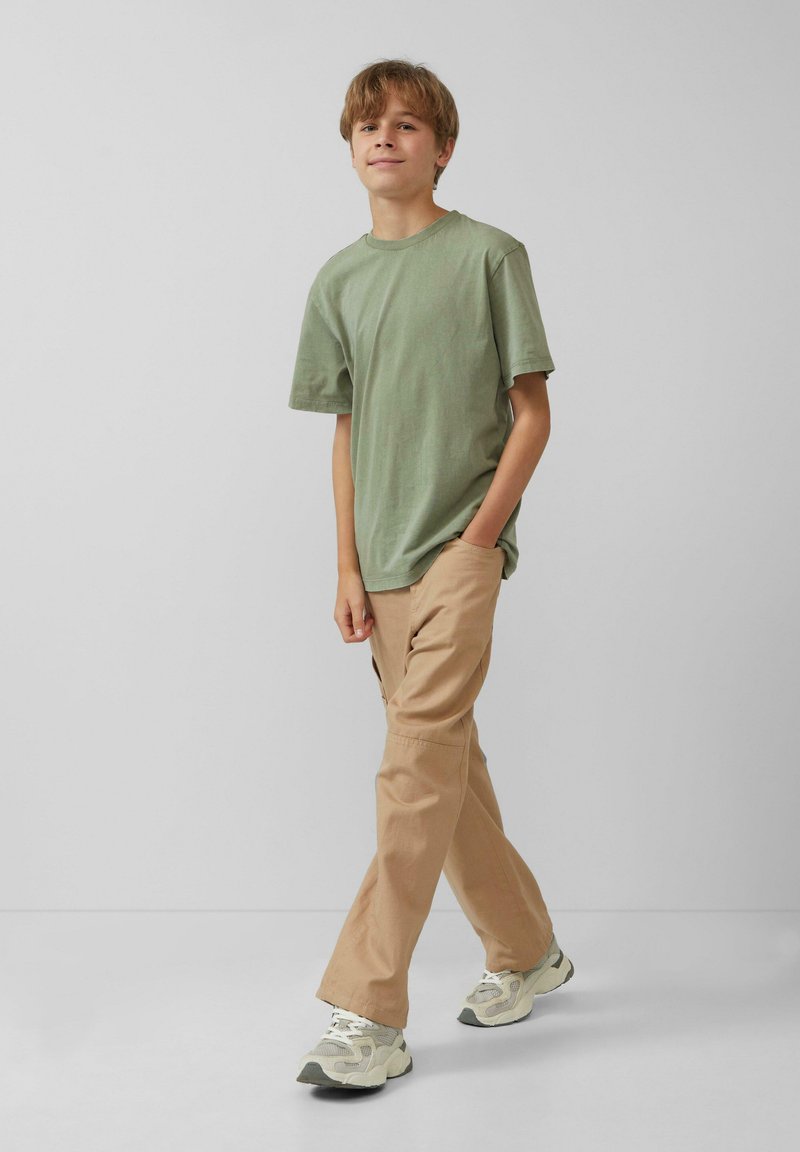 Groene katoenen T-shirt, ontspannen pasvorm, gecombineerd met beige broek. Sneakers met een textuur in grijs en crème ontwerp. Eenvoudige achtergrond.