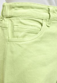 Gros plan sur un pantalon en denim vert clair montrant la poche avant, les coutures et les passants de ceinture.