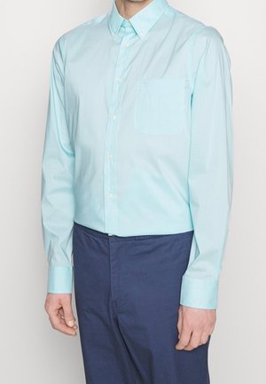 Mężczyzna w jasnoniebieskiej koszuli zapinanej na guziki z kieszenią na piersi i ciemnoniebieskich spodniach, stojący na jednolitym tle.