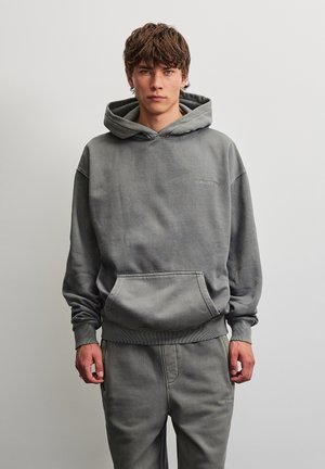Jeune homme debout portant un sweat à capuche gris assorti et un pantalon de jogging, les mains détendues le long du corps, sur un fond clair uni.