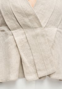 Κοντινή λήψη υφάσματος μπεζ λινό με υφή, με επικαλυπτόμενα φαρδιά πάνελ και ορατή ραφή, φορεμένο πάνω από ανοιχτόχρωμο παντελόνι.