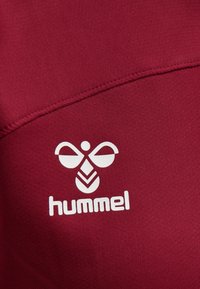 Mørkerødt stof med en glat tekstur har et hvidt Hummel-logo bestående af et stiliseret bi-design over brandnavnet.
