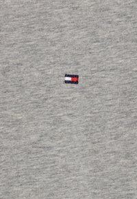 Grå bomullströja med en liten broderad logotyp som har röda, vita och blå ränder, placerad på vänster sida av bröstet.