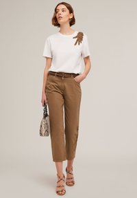 Camiseta de algodón blanca con un diseño texturizado marrón en el hombro, combinada con pantalones cortos marrones y un bolso con estampado.