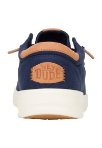 Scarpe in tela blu navy con un accento in pelle color cuoio sul retro, caratterizzate dal logo "HEY DUDE" impresso su una toppa circolare. Suola bianca.