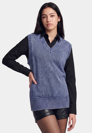 Vrouw die een blauwe mouwloze gebreide vest draagt over een zwart shirt met lange mouwen en zwarte leren korte broek, poserend met één hand op haar heup.