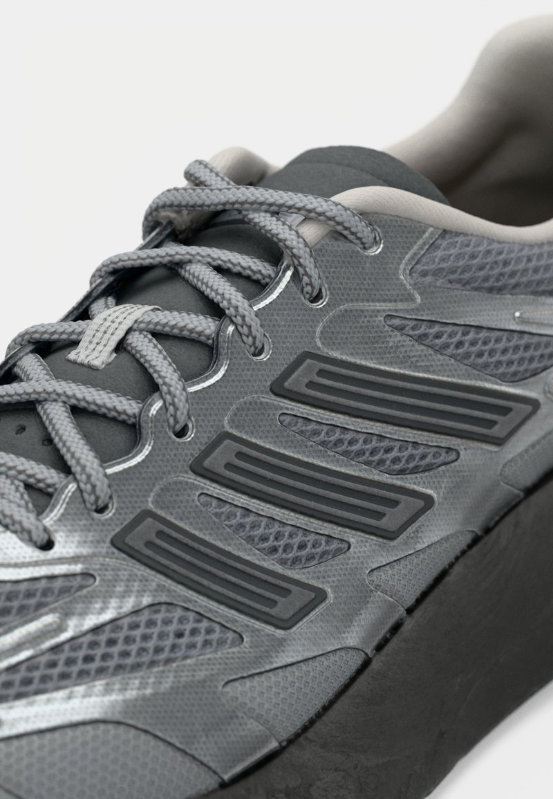 Grauer Sportschuh mit strukturiertem Obermaterial, ausgestattet mit Netzpanels, schwarzen Gummielementen und grauen Schnürsenkeln. Dezente reflexive Details sind vorhanden.