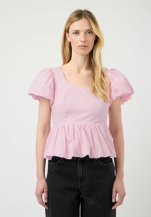 Blond kvinne iført en lyserosa bluse med pufferm og firkantet utringning, og svarte høytlivsjeans, står foran en ensfarget bakgrunn.