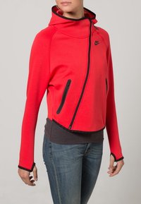 Red zip-up hoodie with a high collar, black accents, and zippered side pockets. Fitted design with long sleeves and a textured fabric.