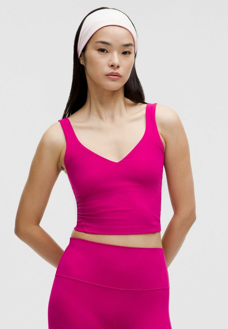 Růžový crop top se zapadajícím střihem, véčkovým výstřihem a širokými ramínky, vyrobený z hladkého materiálu. Modelka má na sobě odpovídající růžové kalhoty s vysokým pasem.