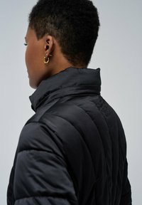 Chaqueta acolchada negra con cuello alto, que presenta un diseño acolchado y una textura suave. Arete de aro dorado visible en el modelo.
