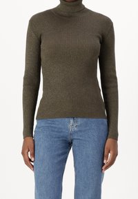 Femme portant un pull en col roulé côtelé vert olive ajusté et un jean taille haute bleu clair, debout avec les bras relâchés le long du corps.