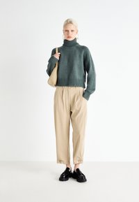 Green knitted turtleneck sweater with ribbed hem, paired with beige pleated trousers and black shoes. Model holds a beige shoulder bag.