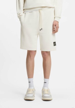 Uomo che indossa pantaloncini color crema con coulisse, calzini bianchi con testo nero e sneakers crema e grigie, in piedi su sfondo bianco.