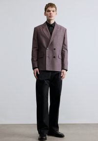 Doppelreihiger Blazer aus hellviolettem Stoff mit strukturiertem Muster, kombiniert mit schwarzem Hemd und Hose, abgerundet mit schwarzen Schuhen.