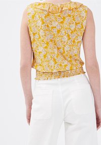 Haut sans manches jaune à motif floral avec encolure froncée et ourlet smocké, porté avec un pantalon taille haute blanc muni de poches arrière.