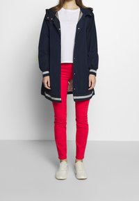 Marineblauwe regenjas met capuchon en zilveren accenten, knoopsluiting en elastische manchetten, in combinatie met slim-fit rode broeken en lichte sneakers.