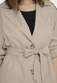 Femme portant un trench-coat beige avec ceinture en tissu nouée et boutons en écaille, par-dessus un haut noir, cadré aux épaules et à la taille.