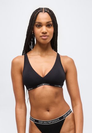Femme aux longs cheveux tressés portant un bralette noir Emporio Armani et une lingerie assortie sur un fond gris uni.