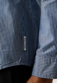 Manșetă și tiv de cămașă stripedă vertical pe alb și albastru, cu eticheta Marc O'Polo aproape de cusătura laterală pe brațul unei persoane.