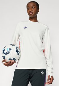 Bílé sportovní tričko s dlouhým rukávem a fialovým logem, spárované s tmavými kalhotami. Míč má texturovaný design s modrými a černými akcenty.