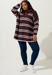 Körsbärsröd stickad tröja med vita ränder, med en halv dragkedja i kragen. Kombinerad med mörkblå jeans och vita sneakers. Ribbad struktur genom hela plagget.