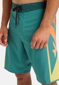 Bañador turquesa con cordón negro, que cuenta con un gran patrón gráfico en naranja y amarillo en el lateral y una textura suave.