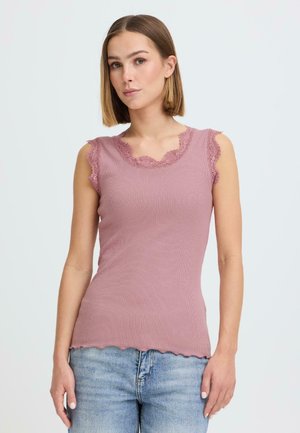 Kvinne med rett, skulderlangt hår som har på seg en ermeløs mauve topp med blonder og lyseblå jeans mot en ensfarget bakgrunn.