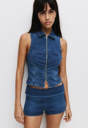 Denim crop vest with a zip front, sleeveless design, and gathered detailing; paired with high-waisted denim shorts in matching blue.