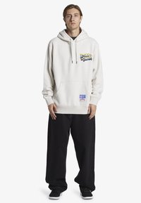 Cream hoodie with a front pocket and graphic print. Paired with loose black pants and black sneakers. Features a woven label at the hem.