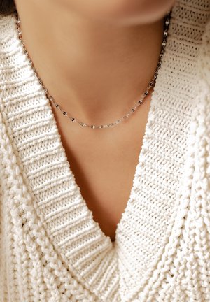 Collar de cadena plateada con pequeñas cuentas negras llevado sobre un jersey de punto grueso con cuello en V de color crema en el cuello de una persona.