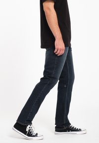 Jeans de mezclilla azul oscuro con corte slim, con costuras sutiles y un logo cerca del dobladillo, combinados con zapatillas negras de caña alta.