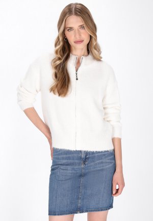Maglione bianco con zip e collo alto, dalla trama morbida e dal taglio loose. Abbinato a una gonna in denim blu, che mostra un outfit casual.