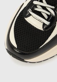 Μαύρο και κρεμ αθλητικό παπούτσι με ανώτερο μέρος από πλέγμα, λείες δερμάτινες λεπτομέρειες, στρογγυλεμένη μύτη και αντίθετα κορδόνια με υφασμένο πάτο.