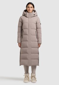 Frau trägt lange, gefütterte Winterjacke in Taupe mit Kapuze, steht aufrecht vor neutralem Hintergrund, beigen Stiefeln und sichtbaren Leggings.