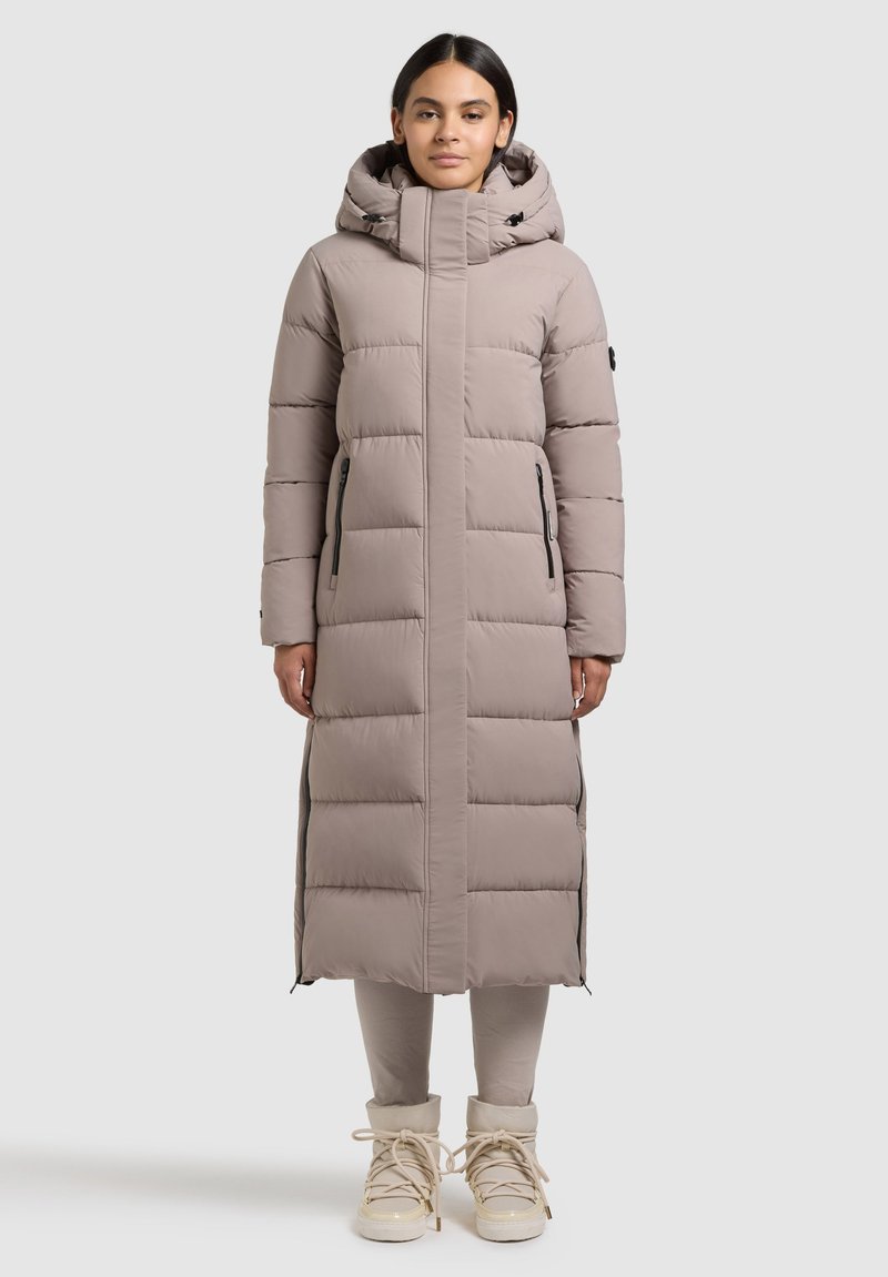 Frau trägt lange, gefütterte Winterjacke in Taupe mit Kapuze, steht aufrecht vor neutralem Hintergrund, beigen Stiefeln und sichtbaren Leggings.