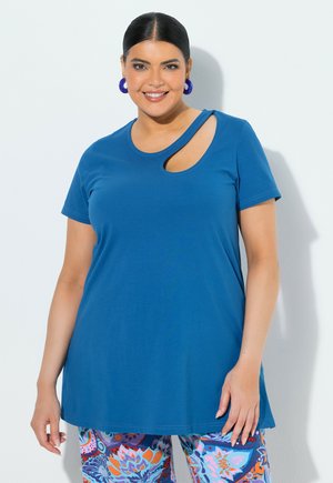 Kvinde iført en blå kortærmet top med et asymmetrisk udskåret mønster og farverige bukser med blomsterprint, smilende foran en ensfarvet baggrund.