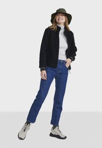 Sort fleecejakke, grå rullekrave, blå jeans, beige sneakers og en olivengrøn bøllehat. Afslappet, løstsiddende outfit med lynlåse og manchetter.